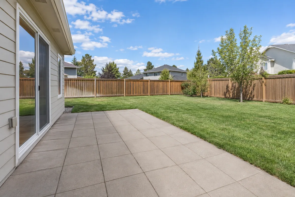 A bare paved patio behind a house, with a lawn and a fence